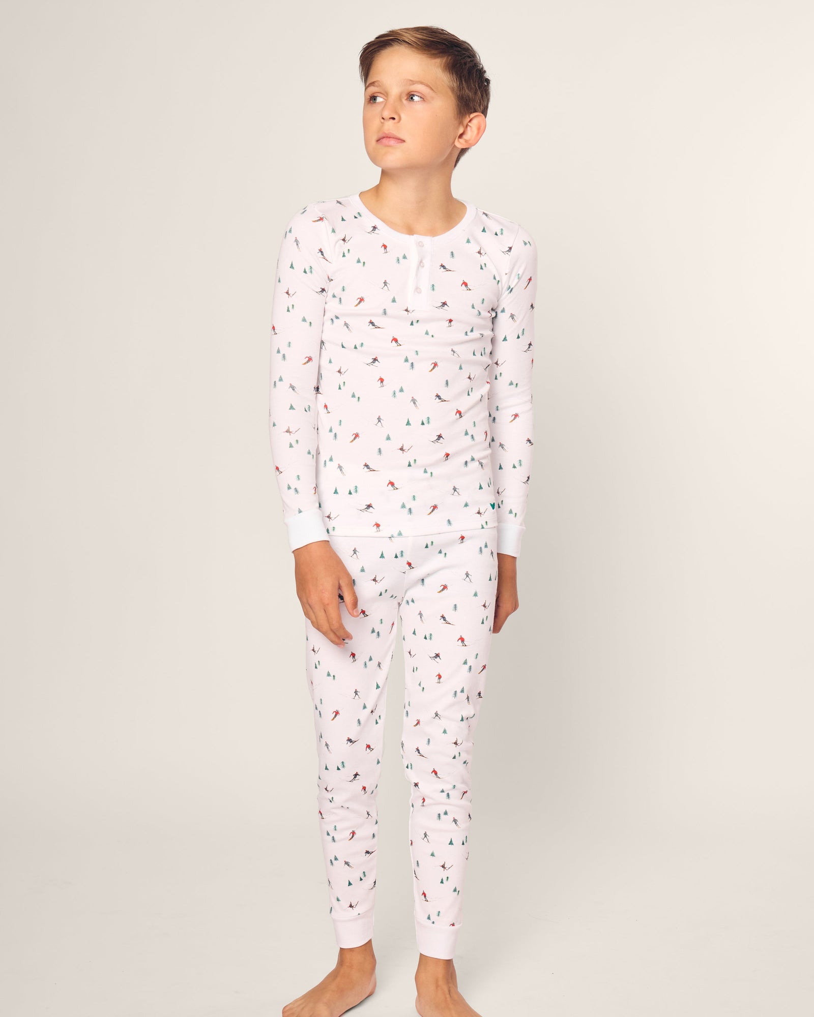 A child stands barefoot against a plain background, wearing Petite Plumes Kids Pima Snug Fit Pajama Set in Apres Ski. The snug long sleeve top features a playful pattern with small trees, tents, and campfires, crafted from soft Pima cotton with a classic button-up style.