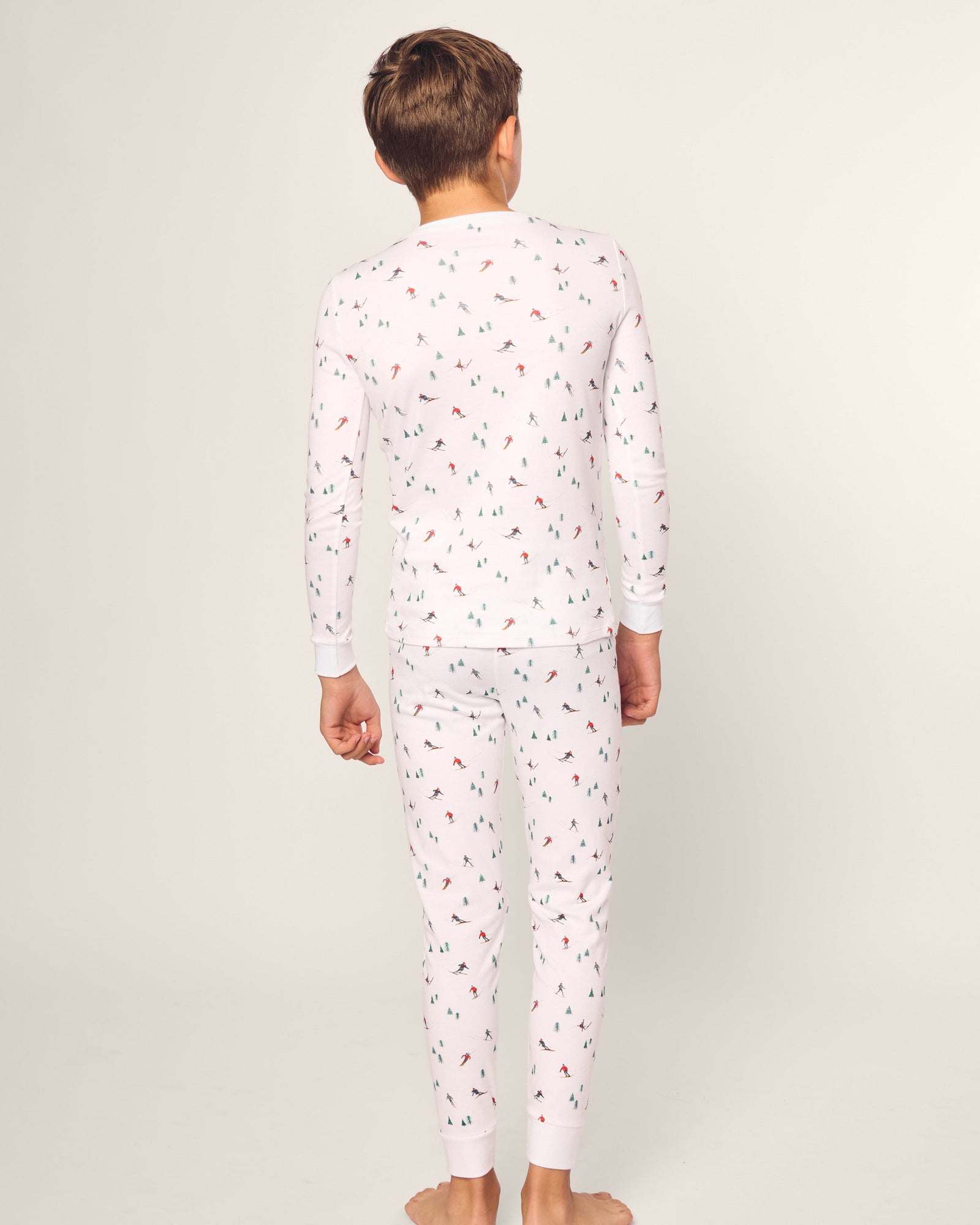 A child stands barefoot, facing away in a snug-fitting Petite Plume Kids Pima Snug Fit Pajama Set in Apres Ski. Made from soft Pima cotton with colorful prints, the pajamas feature long sleeves and pants. The background is a plain, light color.