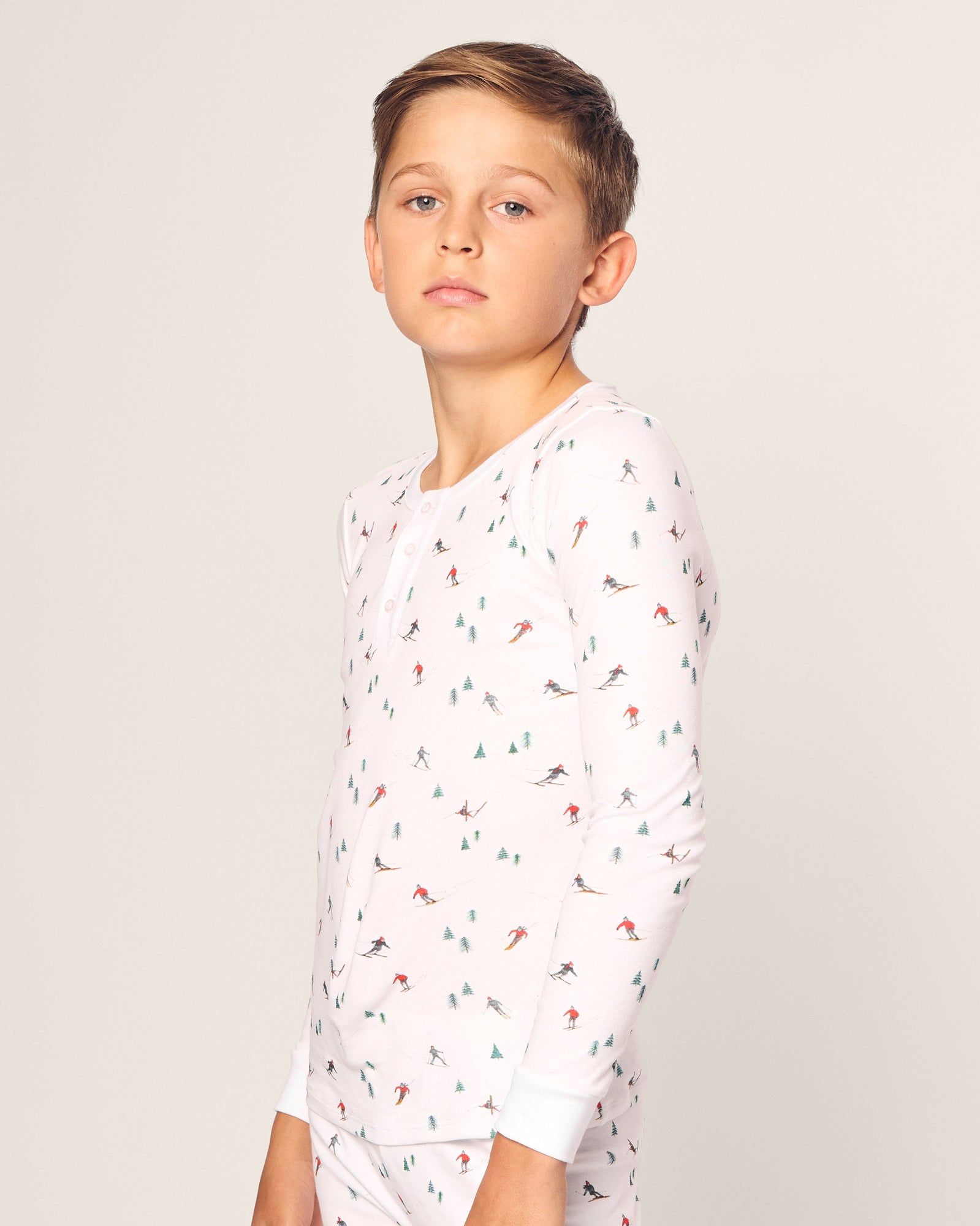A young boy models the Kids Pima Snug Fit Pajama Set in Apres Ski by Petite Plume, featuring soft white Pima cotton with skiing and tree patterns. He stands against a plain background, gazing neutrally at the camera.