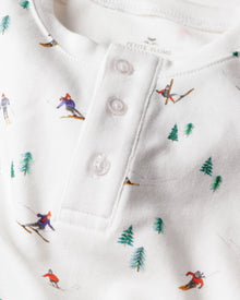 A close-up of Petite Plumes Kids Pima Snug Fit Pajama Set in Apres Ski shows a white long-sleeve top adorned with small skiers and green pine trees, featuring a soft texture and a buttoned placket with three white buttons.