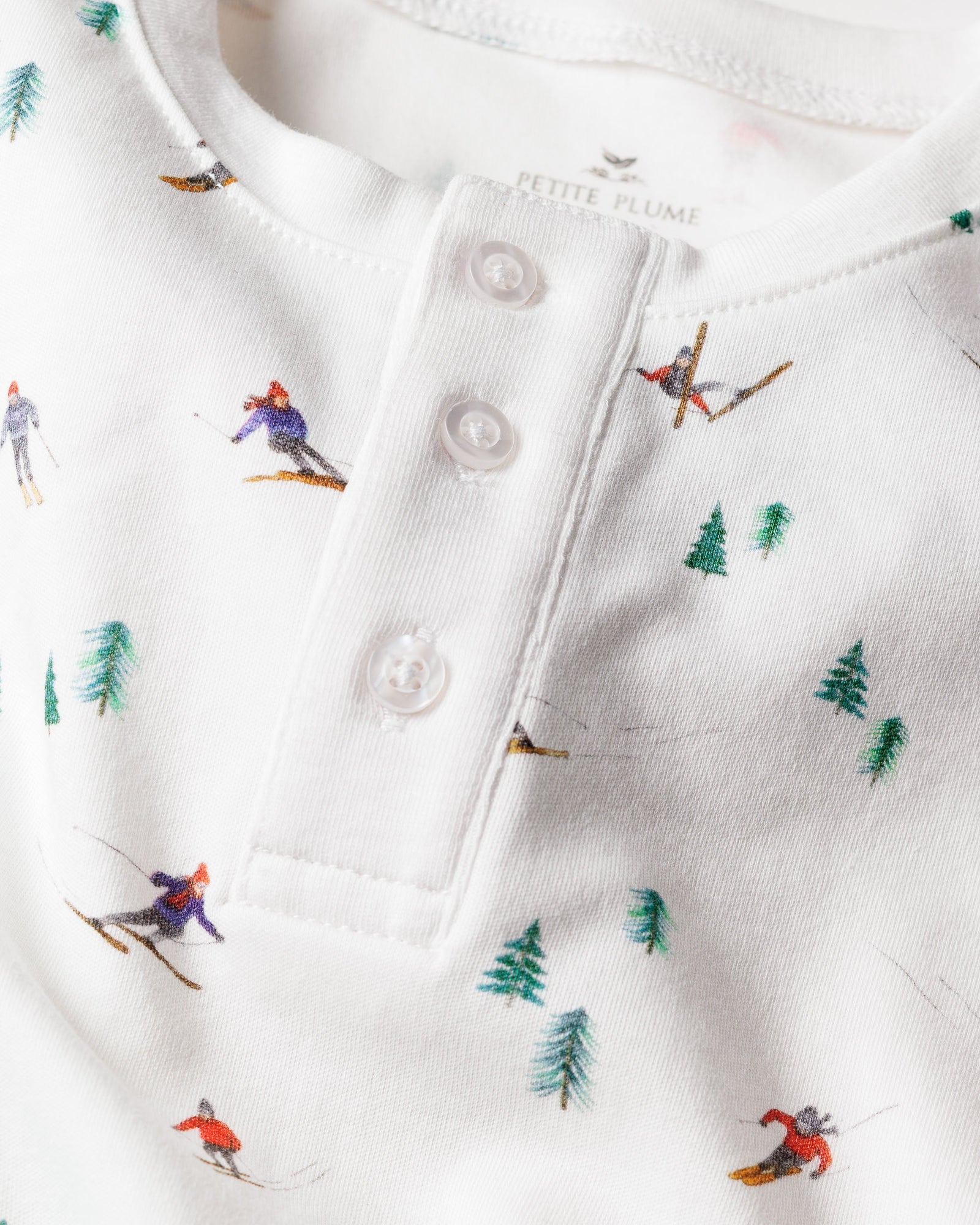 A close-up of Petite Plumes Kids Pima Snug Fit Pajama Set in Apres Ski shows a white long-sleeve top adorned with small skiers and green pine trees, featuring a soft texture and a buttoned placket with three white buttons.