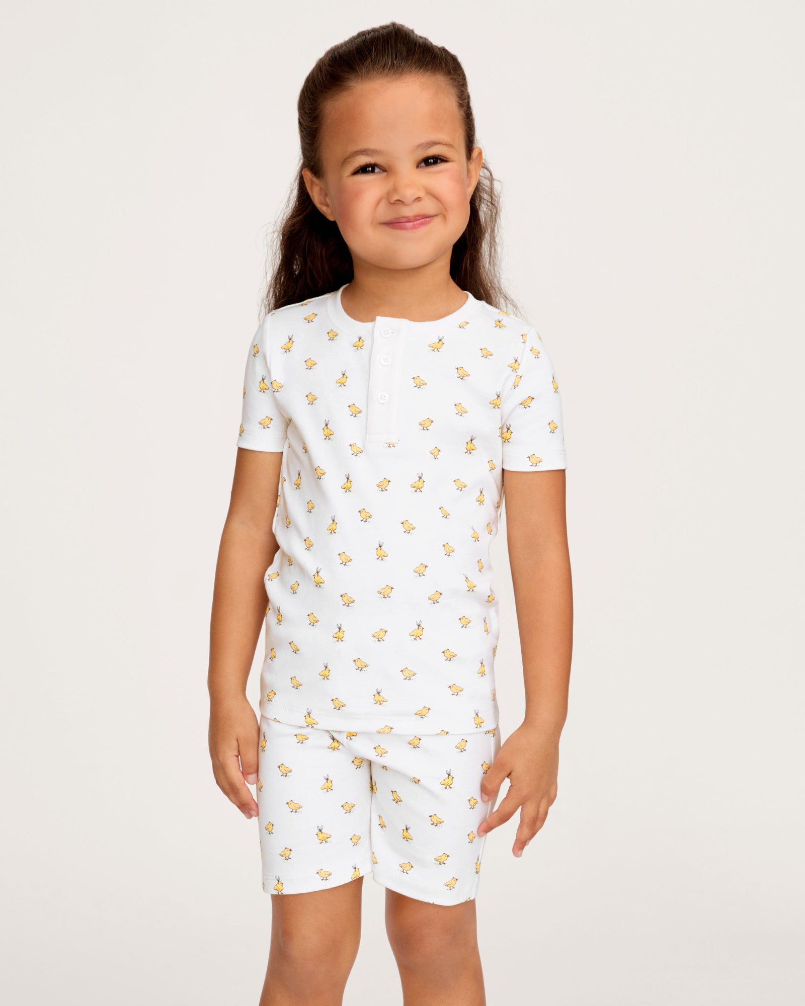 A young child with long brown hair smiles while wearing the Kid's Pima Snug Fit Short Set in Tres Chick, standing against a plain light background.