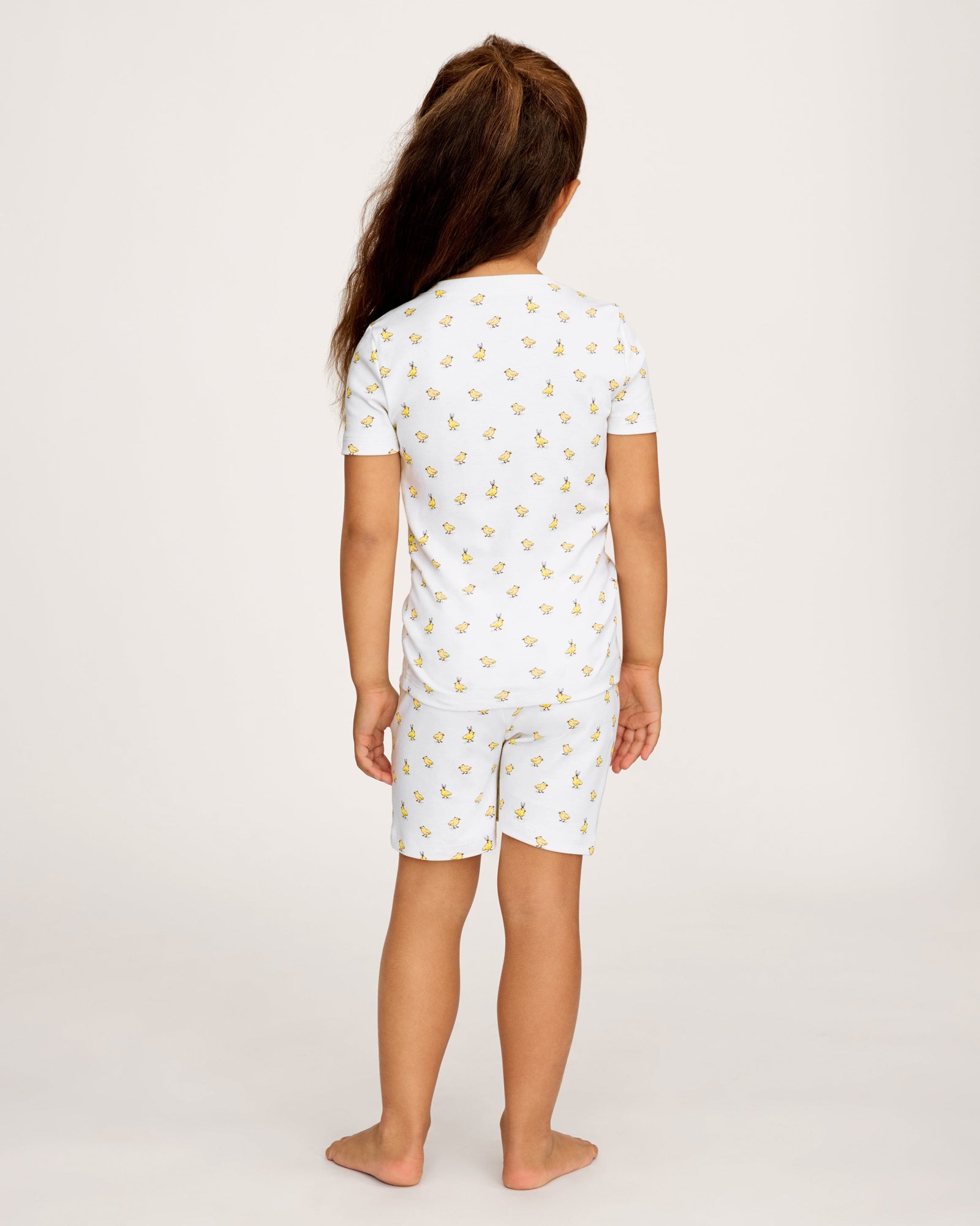 A young child with long brown hair smiles while wearing the Kid's Pima Snug Fit Short Set in Tres Chick, standing against a plain light background.