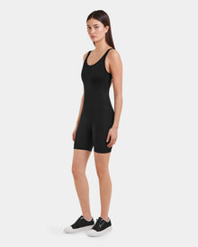A woman with long, straight hair and a neutral expression stands against a white background wearing the Rudsak KAILEY - 8523012 Black Pattern fitted sleeveless bodysuit romper and black sneakers, facing slightly to the side.