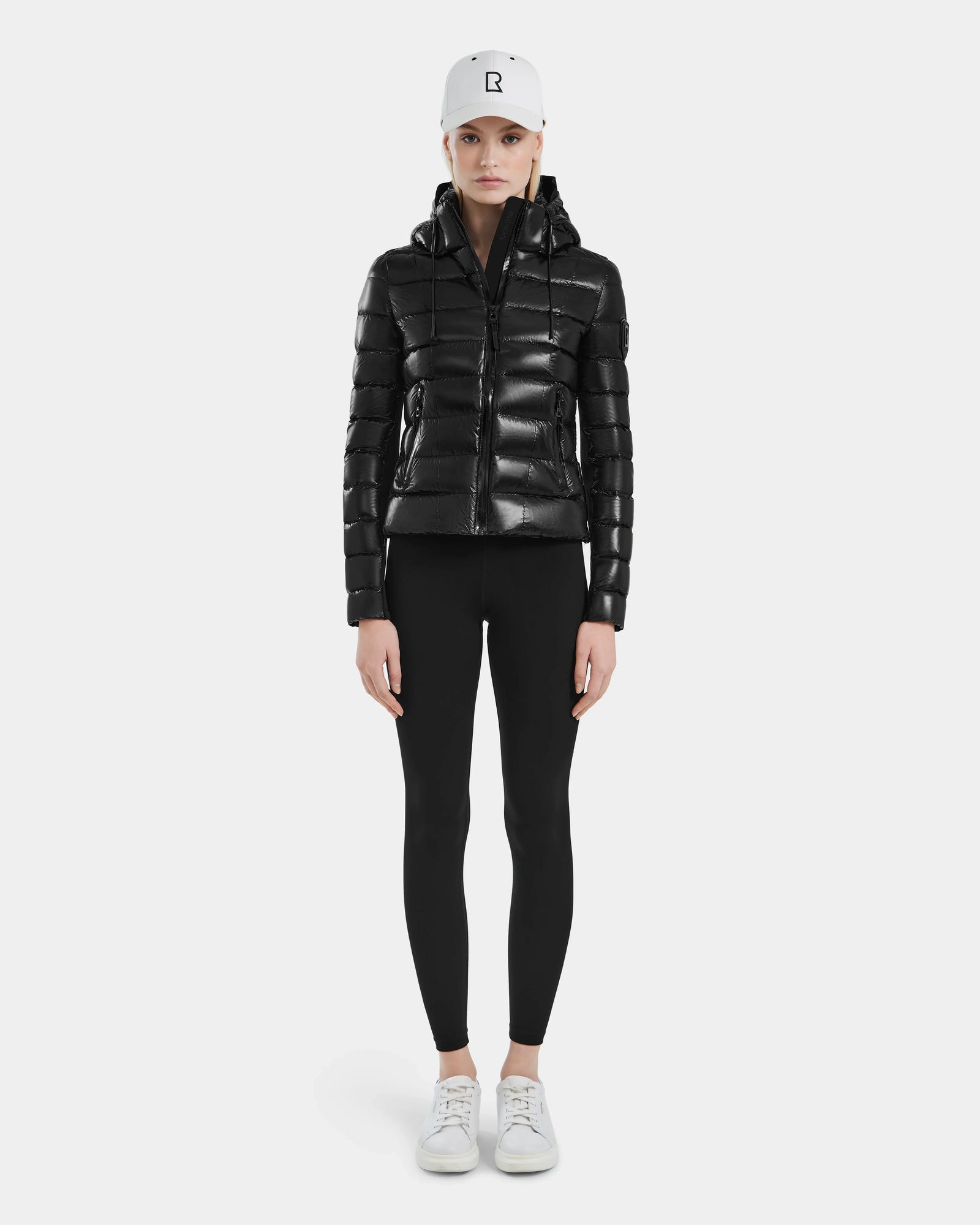 A person stands facing forward in the MELISSA - 8121000 BLACK shiny women’s puffer jacket with RDS certified down, paired with black leggings, white sneakers, and a white baseball cap against a plain light gray background.