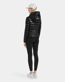 A person stands facing away in the MELISSA - 8121000 BLACK women’s puffer jacket, made from RDS certified down, paired with black leggings, white sneakers, and a white baseball cap against a plain light gray background.