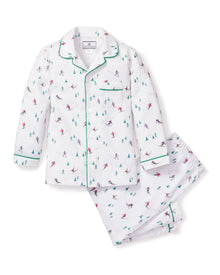 Petite Plumes Kids Flannel Pajama Set in Apres Ski features a white skiing pattern with small figures and trees. The button-down top has green piping on the collar and cuffs, while the matching pants have an elastic waistband. Made from flame-retardant fabric for added safety and comfort.