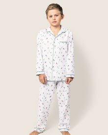 A young boy stands barefoot against a plain background, wearing Petite Plumes cozy Kids Flannel Pajama Set in Apres Ski. These flame-retardant pajamas feature colorful designs, a classic collar, and front buttons for a charming look.
