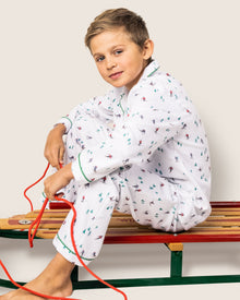 A young boy in Petite Plumes Kids Flannel Pajama Set in Apres Ski sits on a red and green sled, clutching its red rope, looking relaxed against a neutral background.