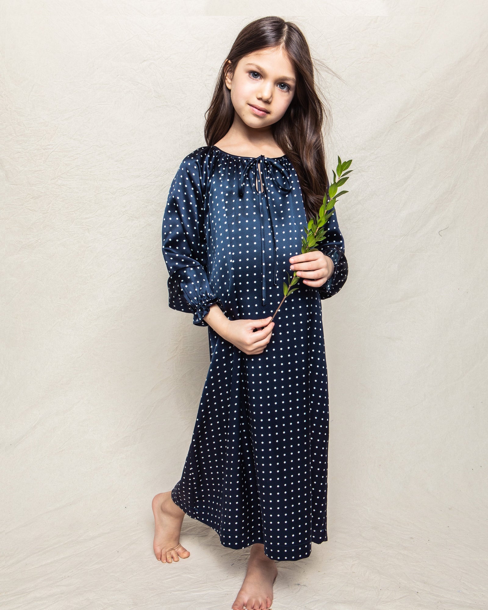 A young girl with long brown hair stands barefoot against a neutral background, wearing a Petite Plume Girls Silk Delphine Nightgown in Navy Polka Dot. She holds a green leafy branch, exuding innocence and charm.