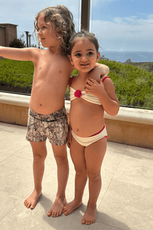 Two children wearing cream and raspberry bikini sets with decorative bindings, standing outdoors on a patio.