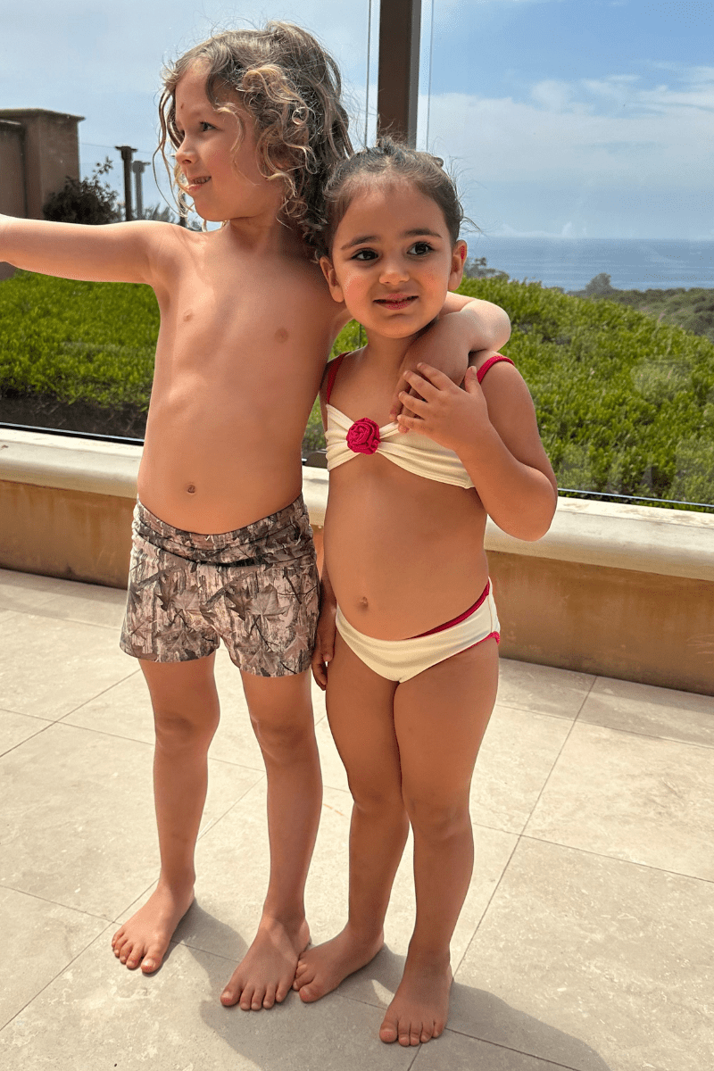 Child wearing tan and brown camouflage print swim trunks on a patio with ocean view.