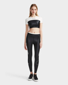 A woman with straight light brown hair stands facing forward, wearing Rudsak’s LILIANE - 8524018 Black women’s fitted crop top and matching high-waisted black leggings with white stripes, paired with black sneakers, against a plain light gray background.