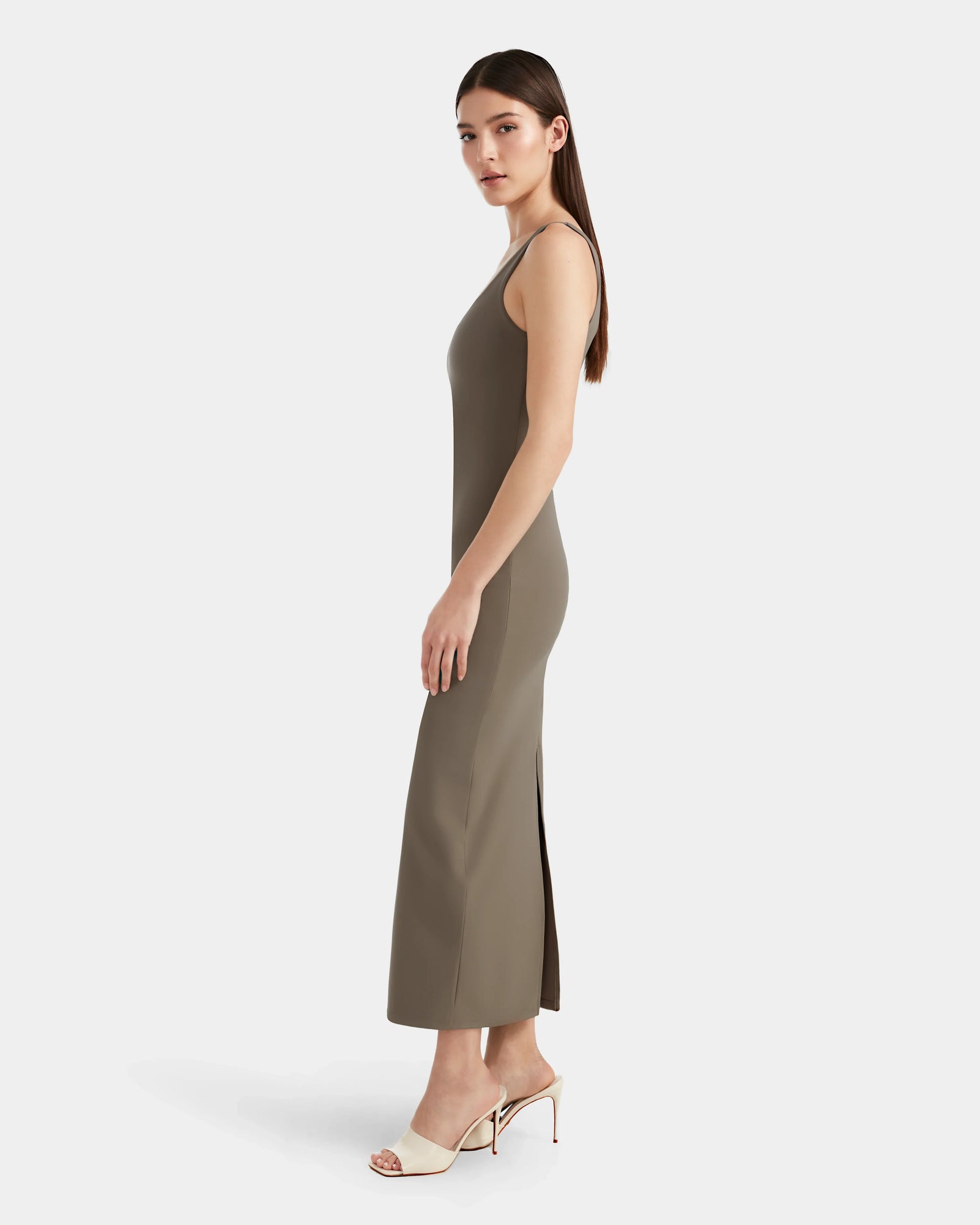 A woman with long straight hair stands sideways, showcasing Rudsaks WANDA - 8524035 Cement maxi dress with a side slit and white heeled sandals, exuding contemporary elegance against a plain white background.