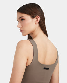 A woman with straight brown hair models the Rudsak WANDA - 8524035 Cement sleeveless stretch performance top, shown from the back and looking over her shoulder against a plain light background.