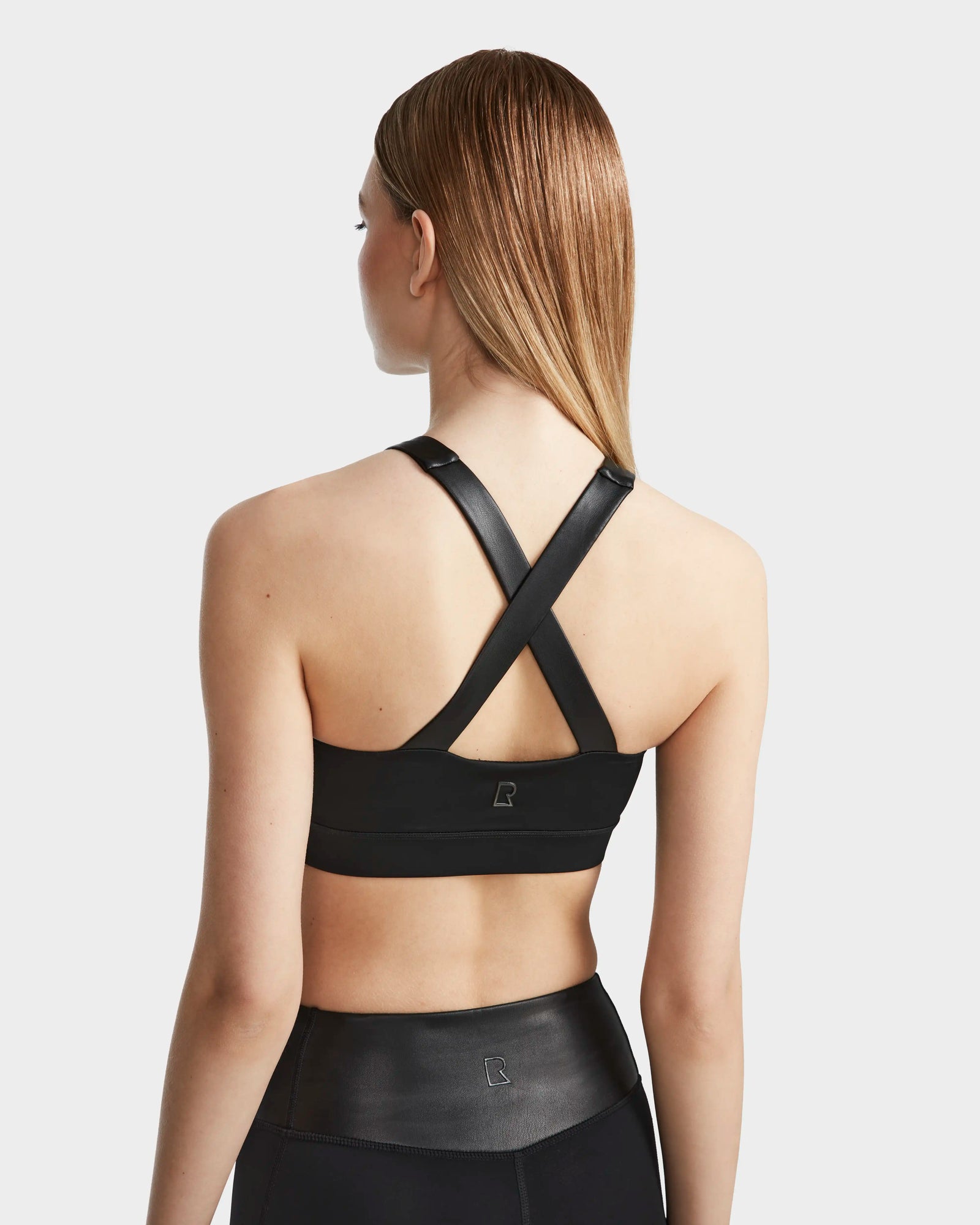 A woman with straight, light brown hair models the RUDSAK ARYA - 8523002 BLACK sports bra, featuring crisscross straps and high-waisted black leggings, both in MVMT+ fabric, shown from the back against a plain light background.