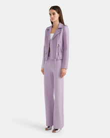 A woman stands against a white background wearing the Rudsak MERGO - 8124034 Lilac Foam pantsuit with a lambskin leather jacket, over a white top and silver open-toe heels.