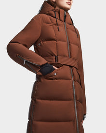A person wearing the RUDSAK CALI - 8122502 COHIBA women’s brown belted winter coat with RDS-certified down, a hood, and zippered details poses with one hand in the pocket against a plain background.