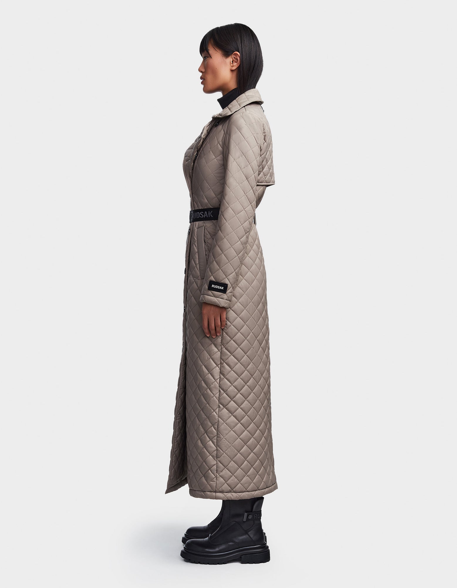 A woman in profile wears the Rudsak DAISY - 8123580 Cement quilted maxi trench coat with a belted waist and black boots. Her straight dark hair contrasts with the plain white background.