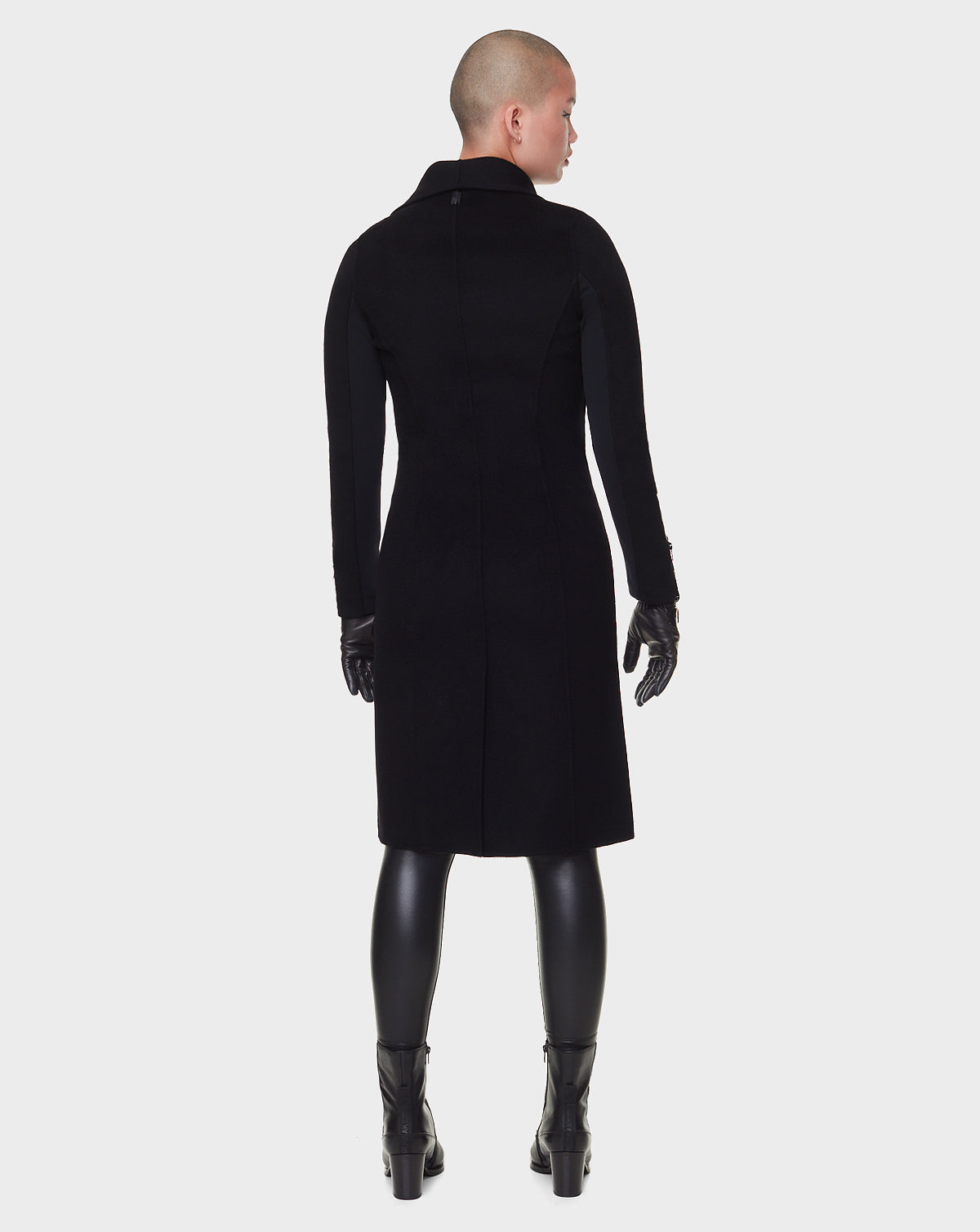 A person with a shaved head models the RUDSAK CHAY - 8122506 BLACK long black wool coat, paired with black gloves, leggings, and boots, standing against a plain white background.