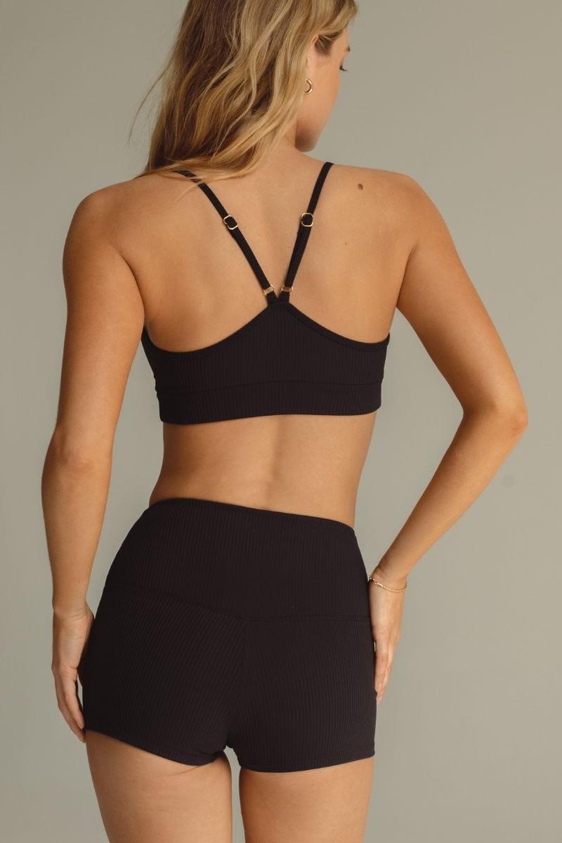 Woman wearing black ribbed micro shorts and matching crop top with adjustable straps, shown from back.