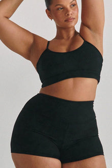 Person wearing black ribbed crop top and matching black micro shorts against neutral background.