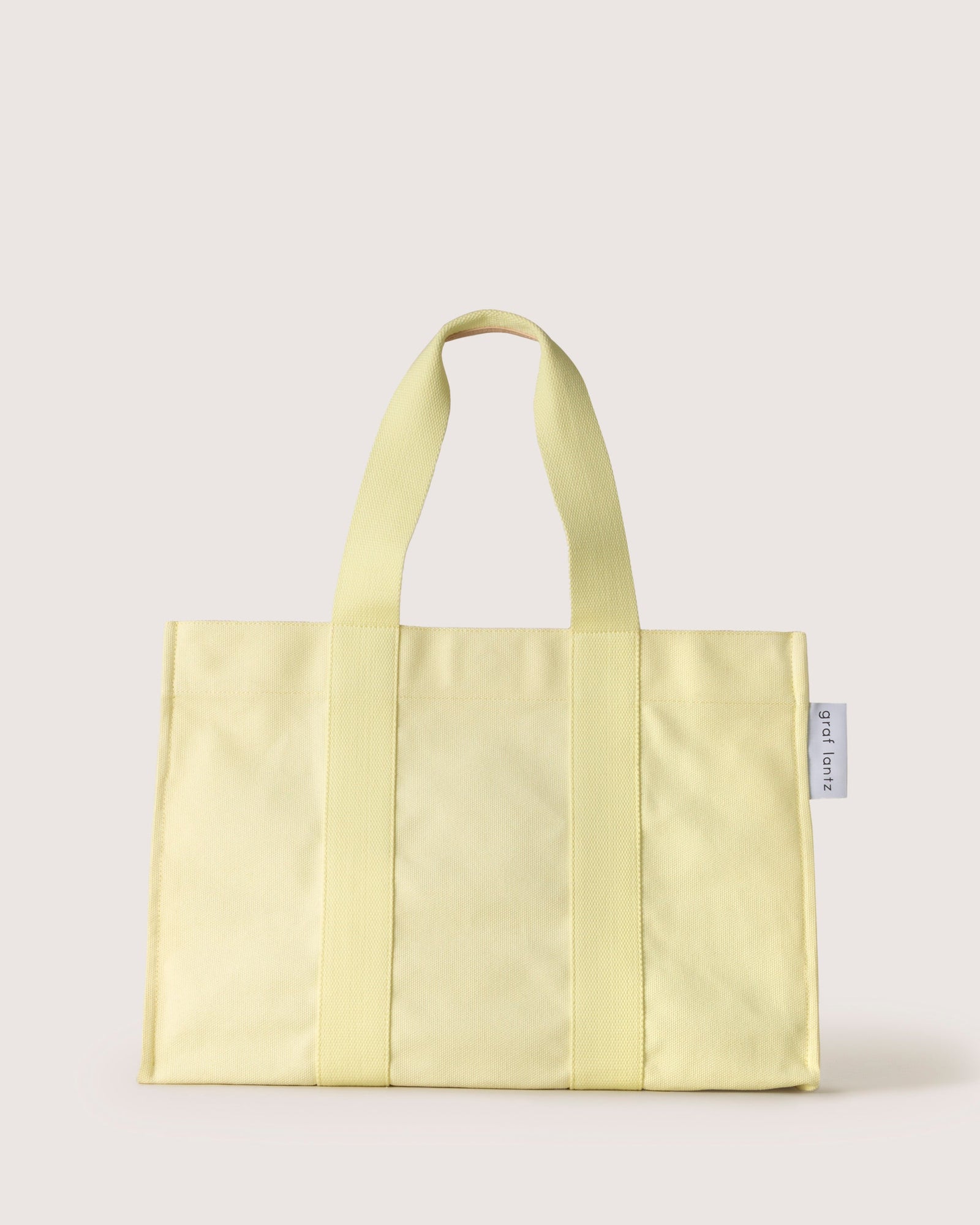 A vibrant yellow graf lantz cotton tote bag, featuring sturdy handles and visible stitching lines, stands upright against a plain light background. Perfect for beach days, it includes a small white tag with text on one side.