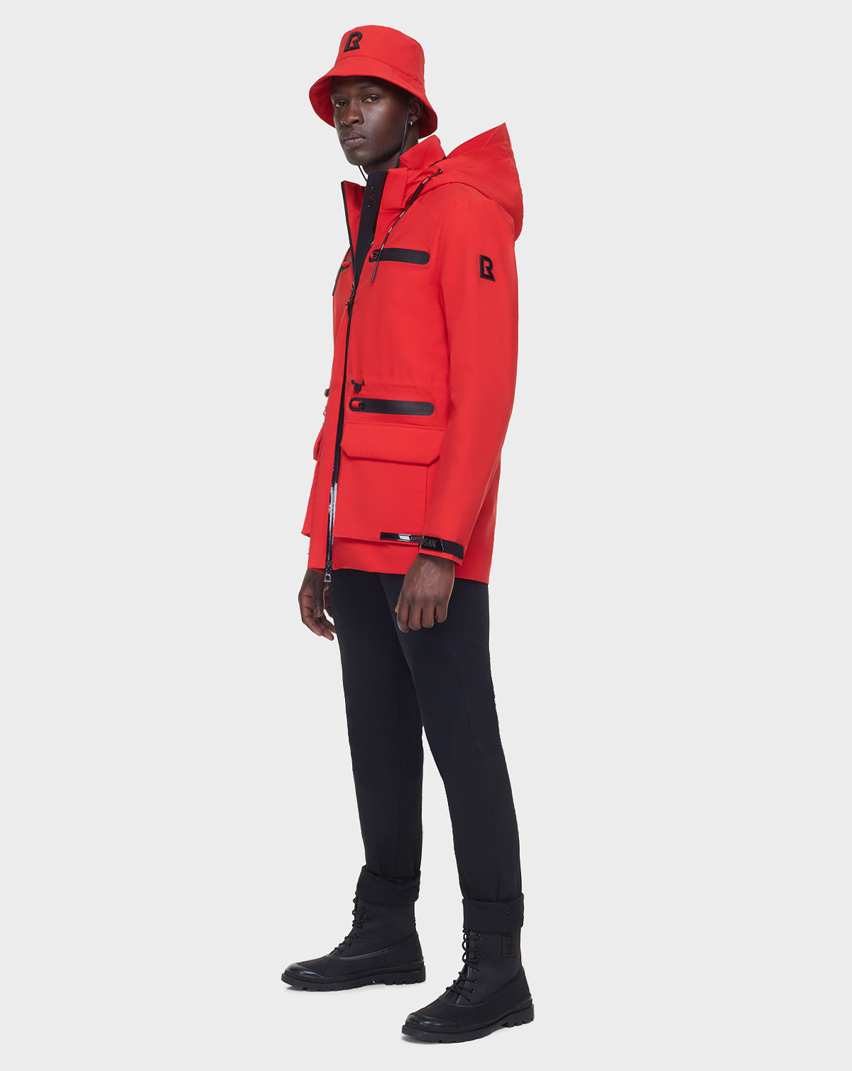 Someone stands against a plain white background wearing the RUDSAK JACY - 6122000 POPPY RED raincoat with a matching red bucket hat, paired with black pants and black boots.